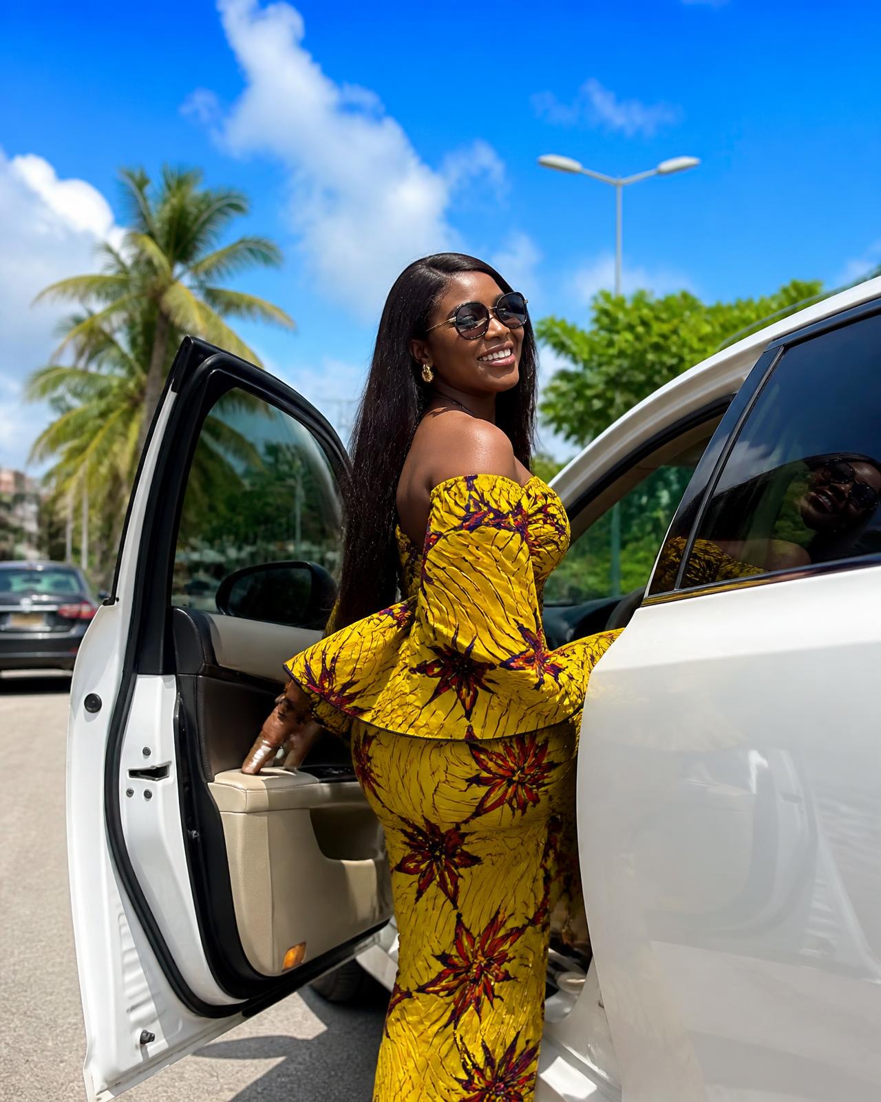 A smiling rider in a bright yellow print dress stepping into a clean white VOYA car on a sunny Lagos street